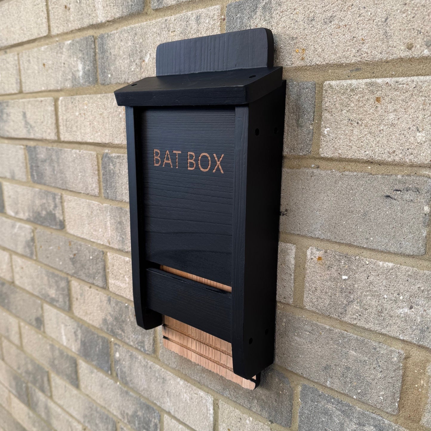 A black wooden bat nesting box mounted on a brick wall, with the words 'BAT BOX' etched on the front.