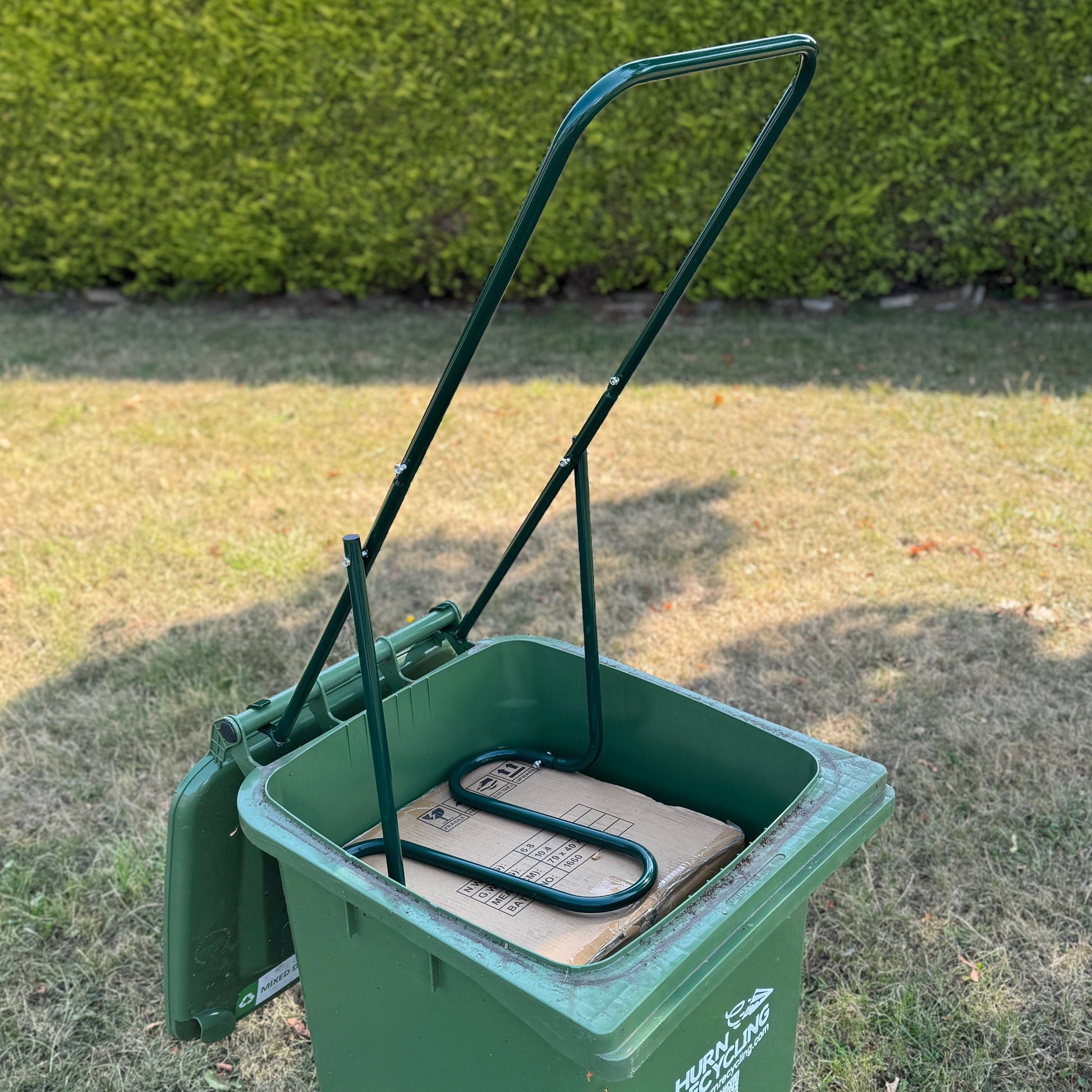 Green wheelie bin with a handle on grass, containing a cardboard box.
