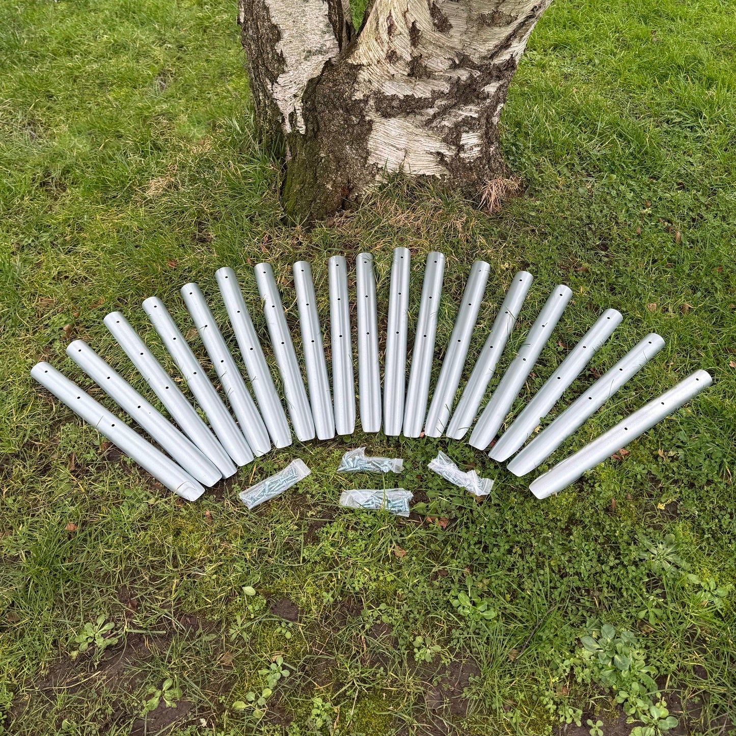A collection of 20 galvanised steel stakes arranged on the grass in front of a tree, with holes for screws and three screws lying on the grass.