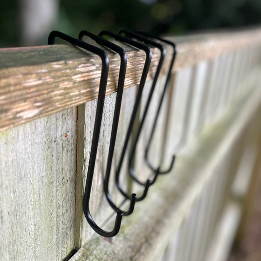 A set of black painted steel hooks designed to be slots over the top of fence panels, pictured against a wooden fence background.