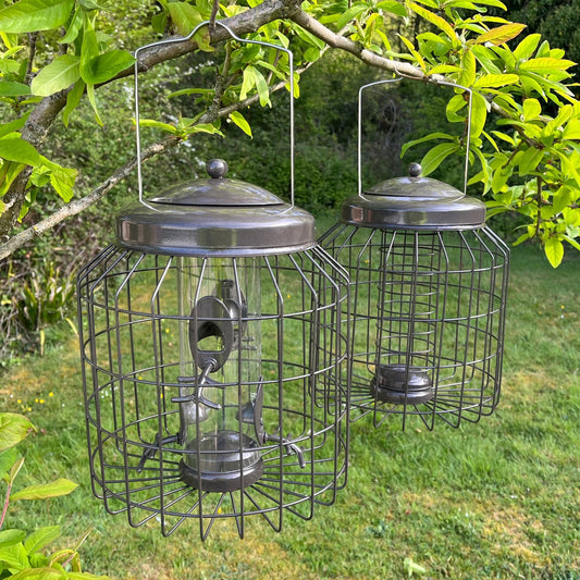Two cylindrical squirrel proof bird feeders with grid-like patterns and hanging hooks, positioned among green foliage.