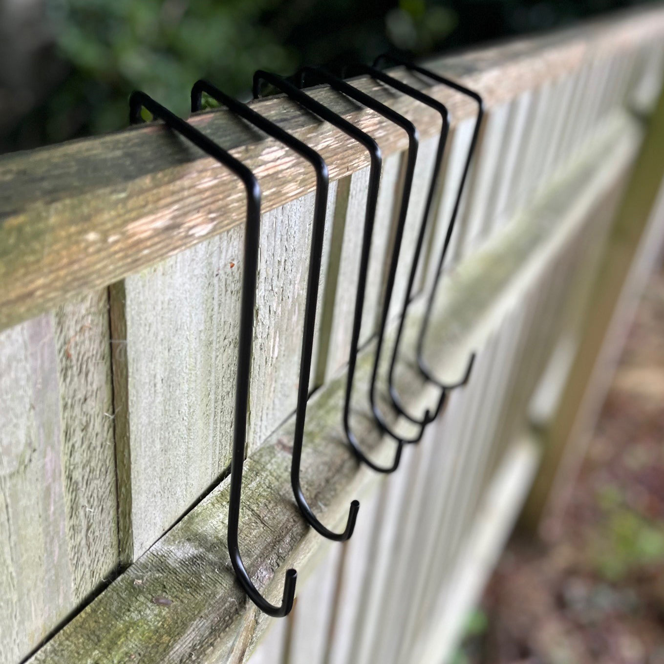 A set of black painted steel hooks slots on a wooden fence panel, designed for hanging planters or decorations.