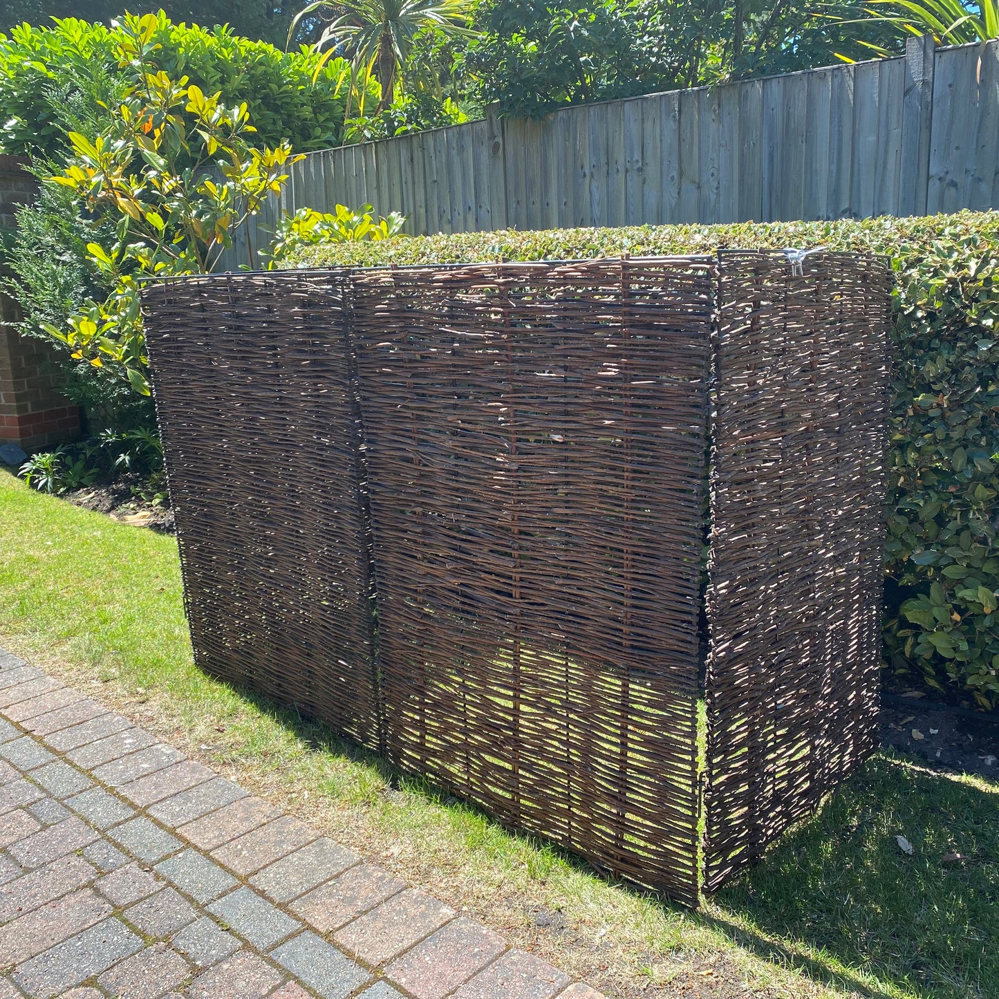 A willow wheelie bin screen covering three plastic bins, with a hedge in the background.