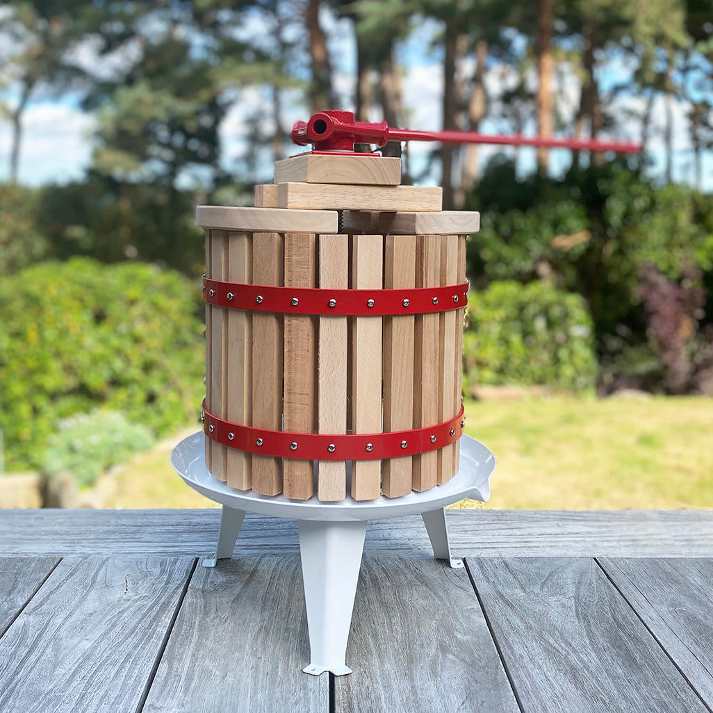 A traditional styled 12 litre fruit press made of wood with a steel base plate and a red straining bag, placed on a wooden surface with a blurred natural background.