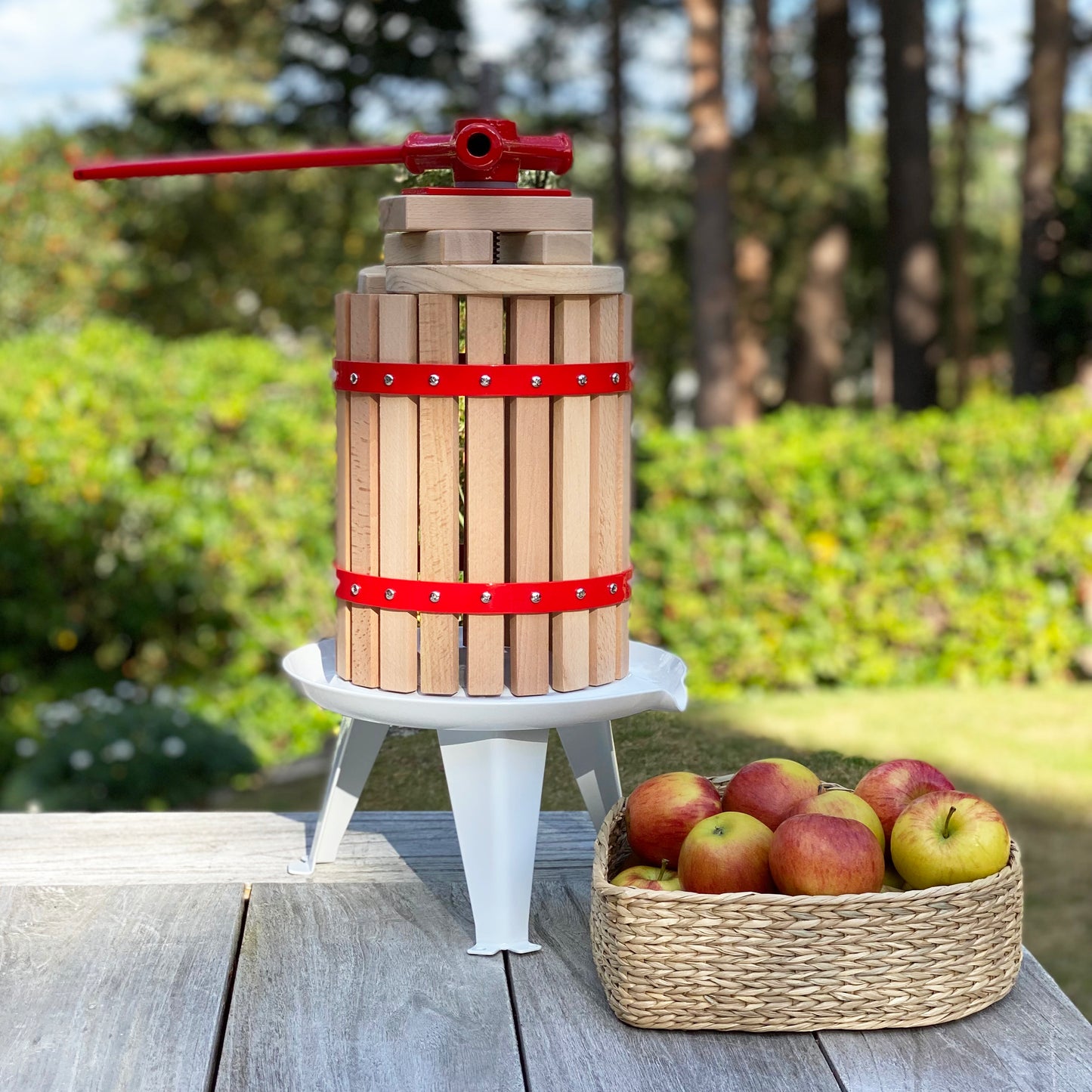 A traditional styled 6 litre fruit press made of wood with a steel base plate, featuring a red pressing handle and a straining bag, alongside a basket of apples.