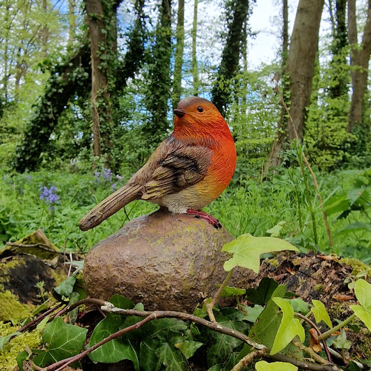 A resin garden ornament depicting a red robin bird sitting on a brown stone, placed in a natural outdoor setting with green foliage in the background.