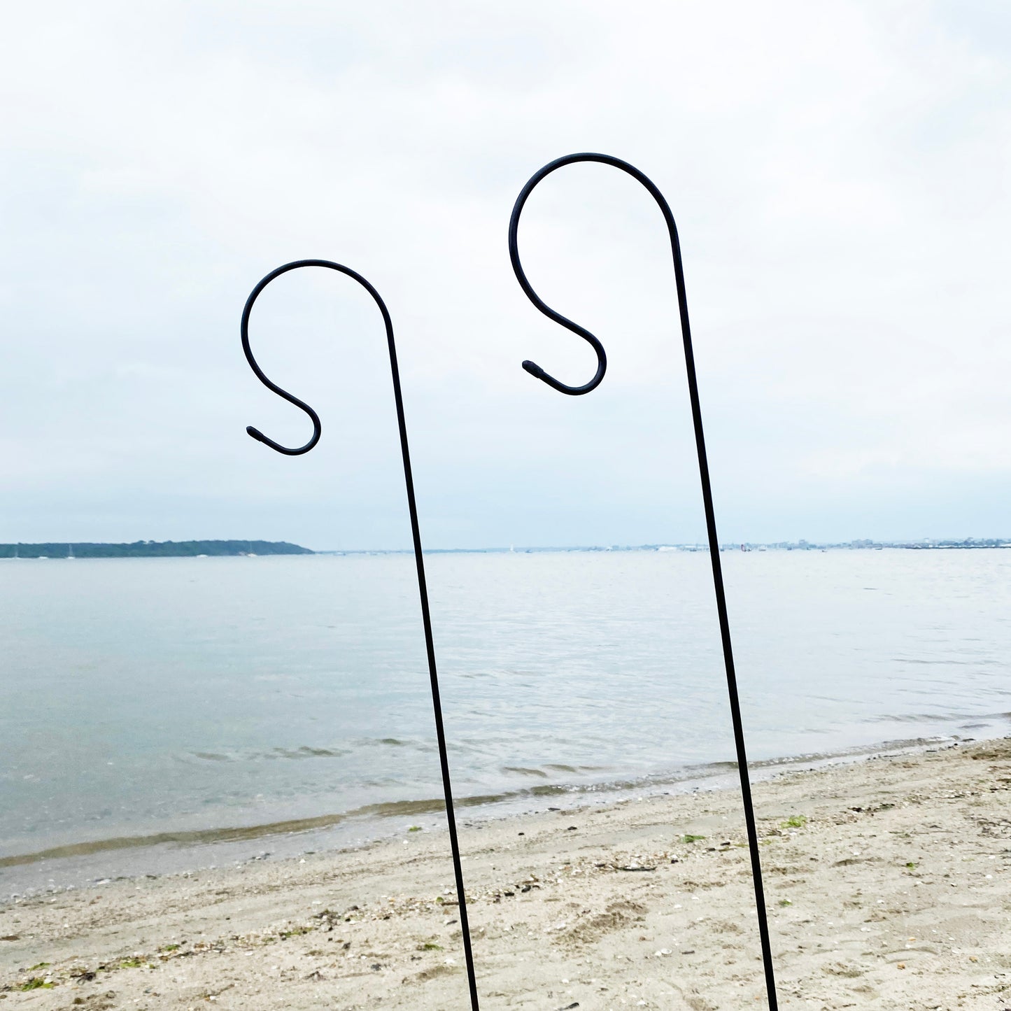 Two black metal garden border hooks standing upright on a sandy surface with a body of water and sky in the background.