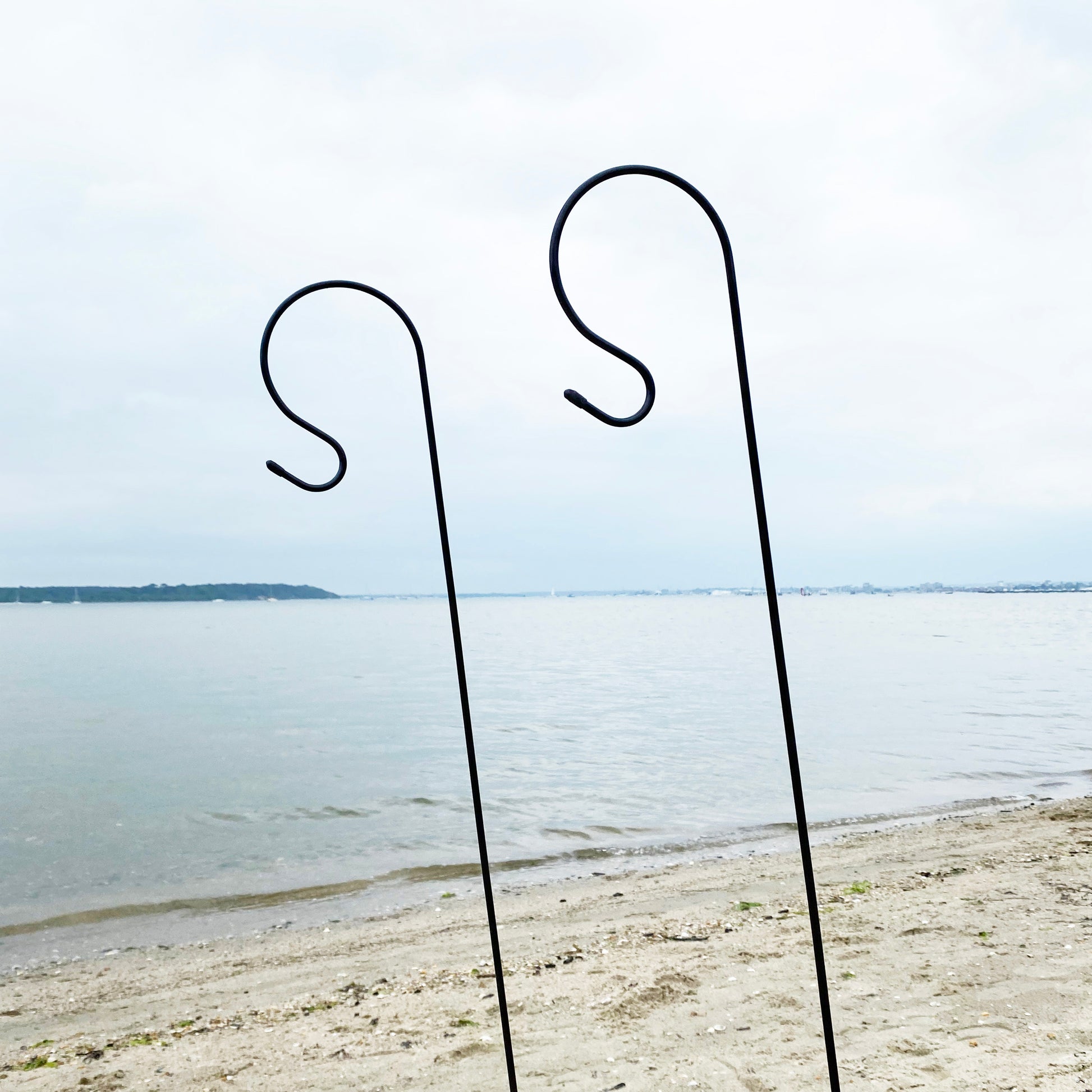Two black metal garden border hooks standing upright on a sandy surface with a body of water and sky in the background.