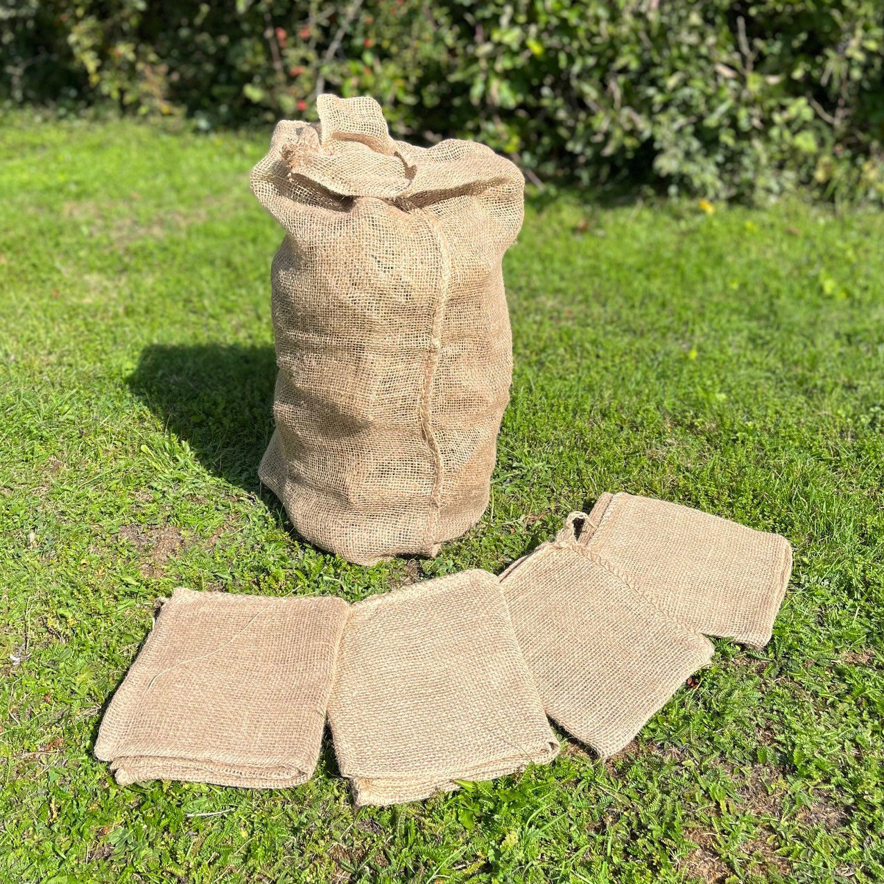 A pack of 5 beige jute potato storage sacks placed on grass, with one sack upright and the others folded.