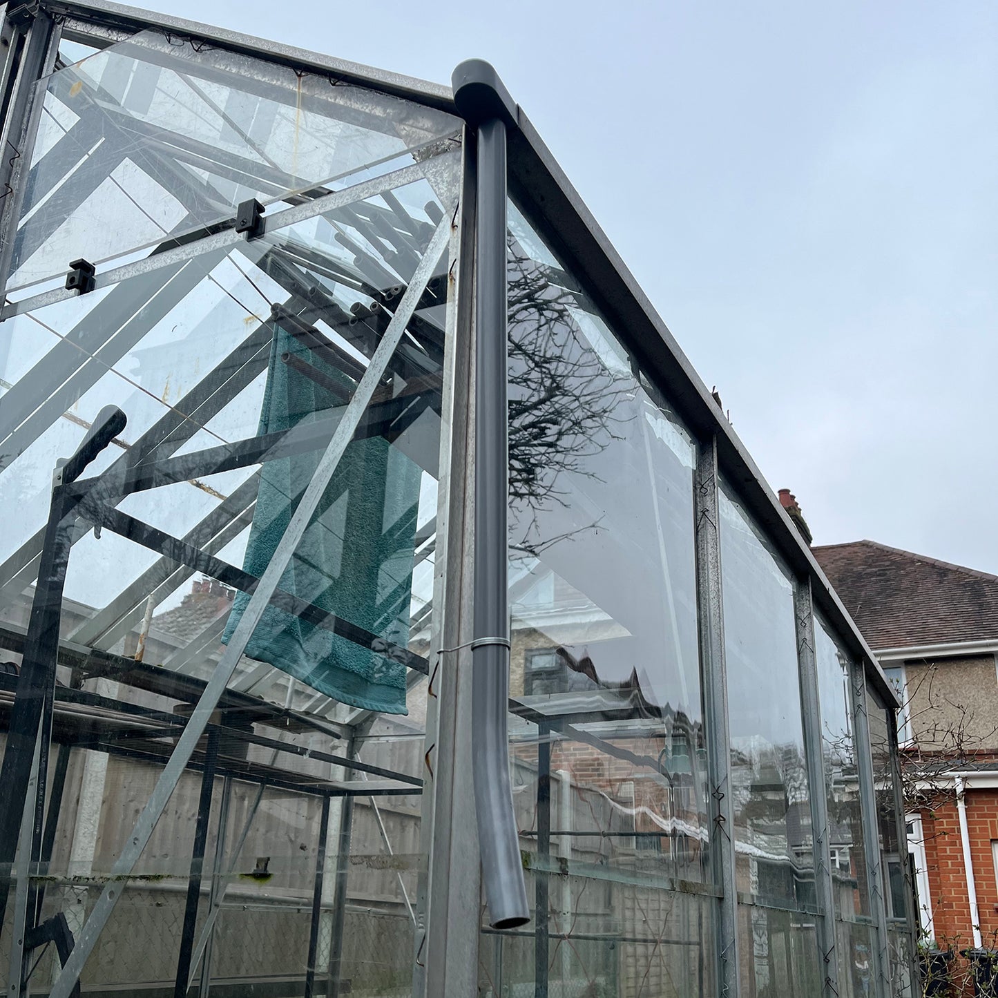 Greenhouse guttering rainwater kit consisting of a grey metal downpipe attached to the end of a clear greenhouse gutter.