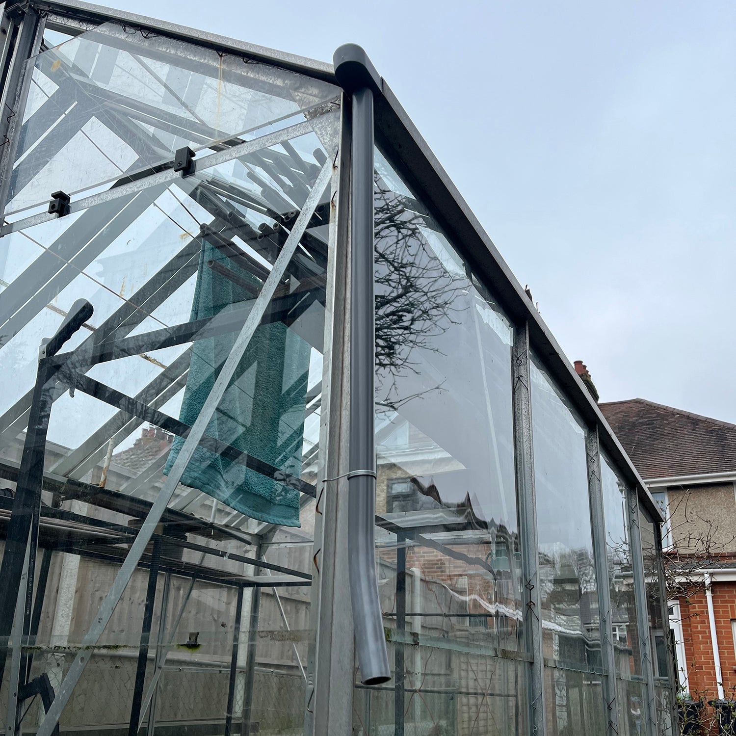 Greenhouse guttering rainwater kit consisting of a grey metal downpipe attached to the end of a clear greenhouse gutter.