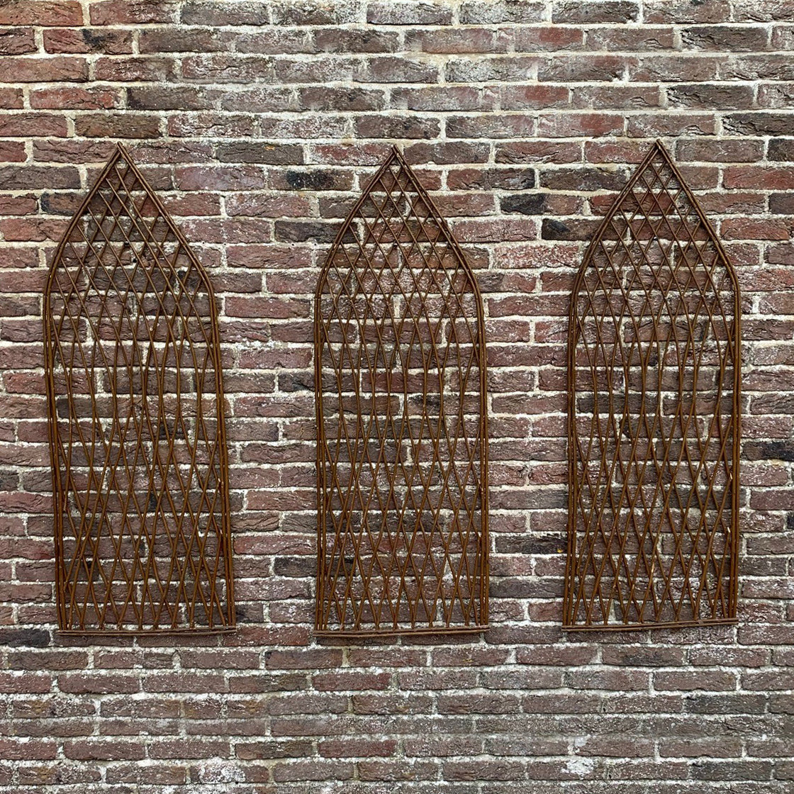 A set of three brown willow trellises with gothic styled tops, mounted on a brick wall.