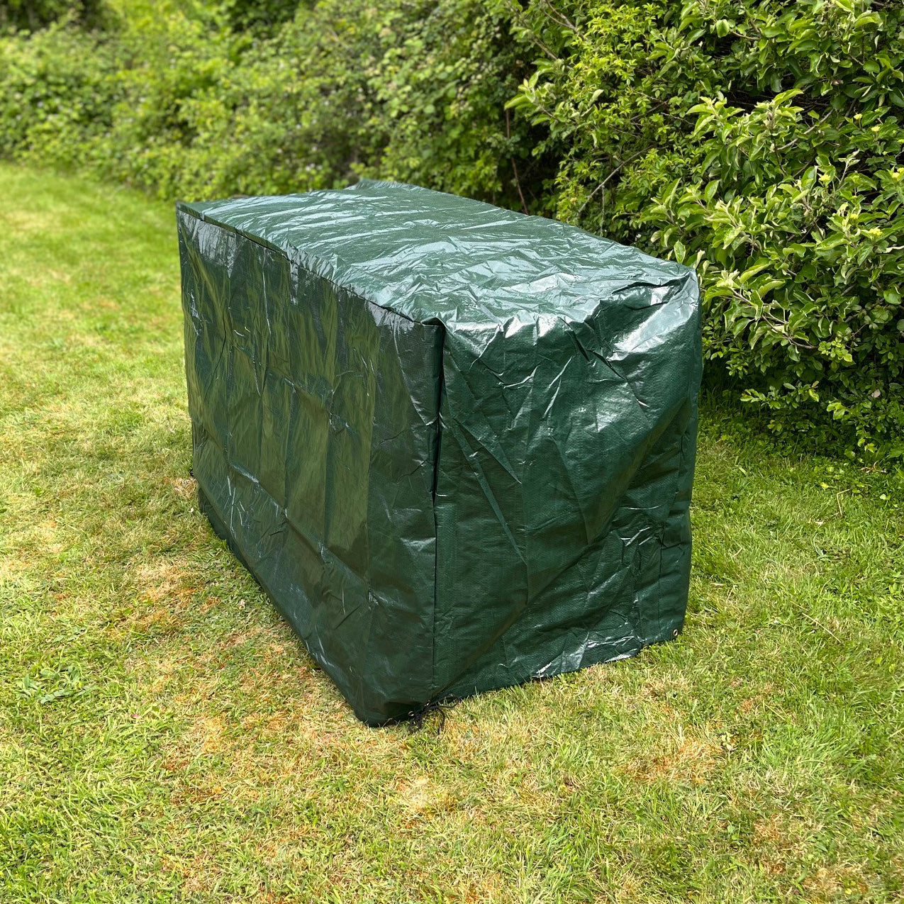 A green waterproof bench cover over a garden bench, placed on grass with foliage in the background.