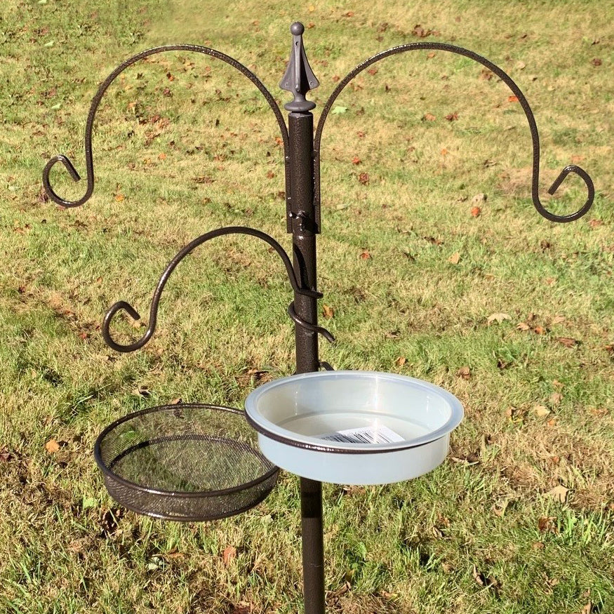 A metal bird feeding station with a mealworm tray and water dish, standing on grass.