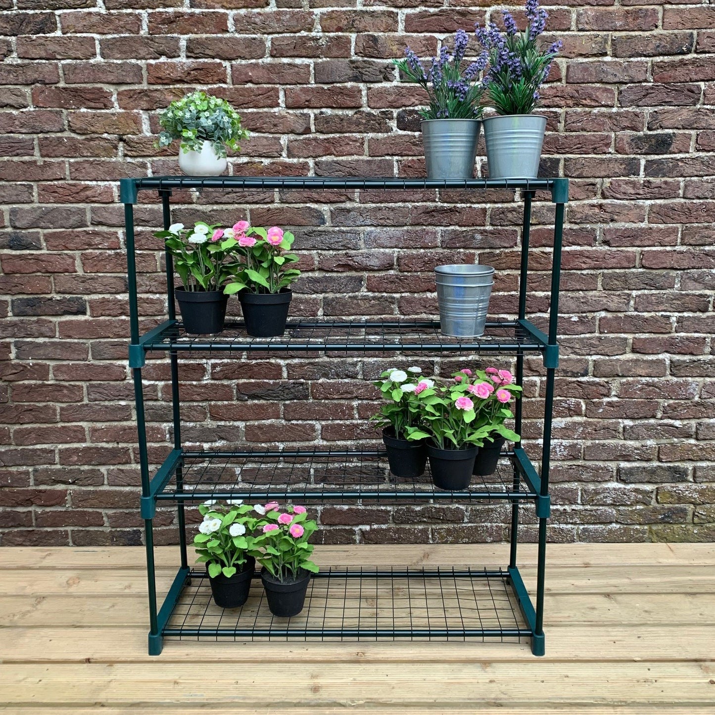 A 4-tier green shelving unit with mesh panels, containing several potted plants on each shelf, placed against a brick wall on a wooden floor.