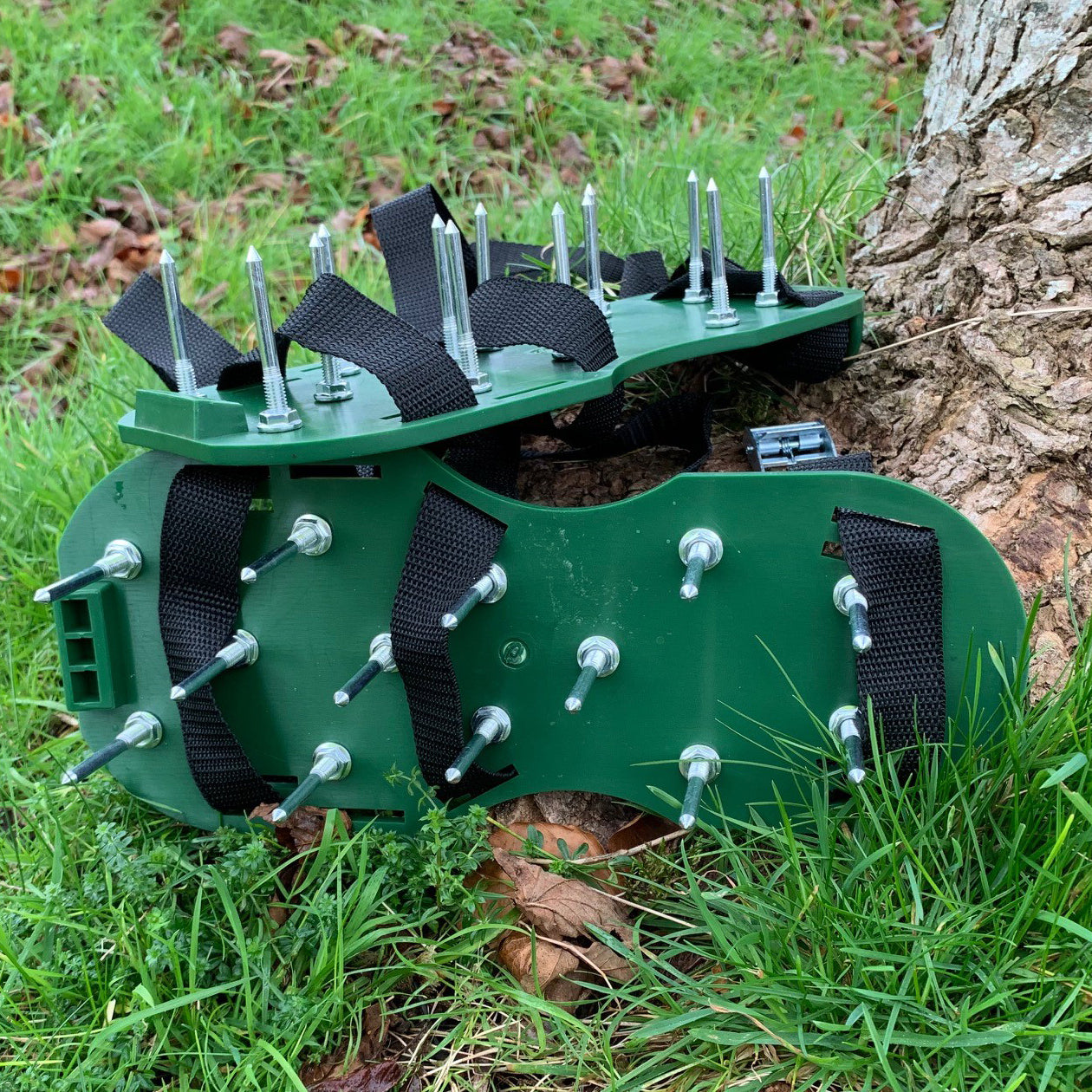 A pair of green spiker shoes designed for lawn aeration, with spikes attached to the soles, placed on grass next to a tree.