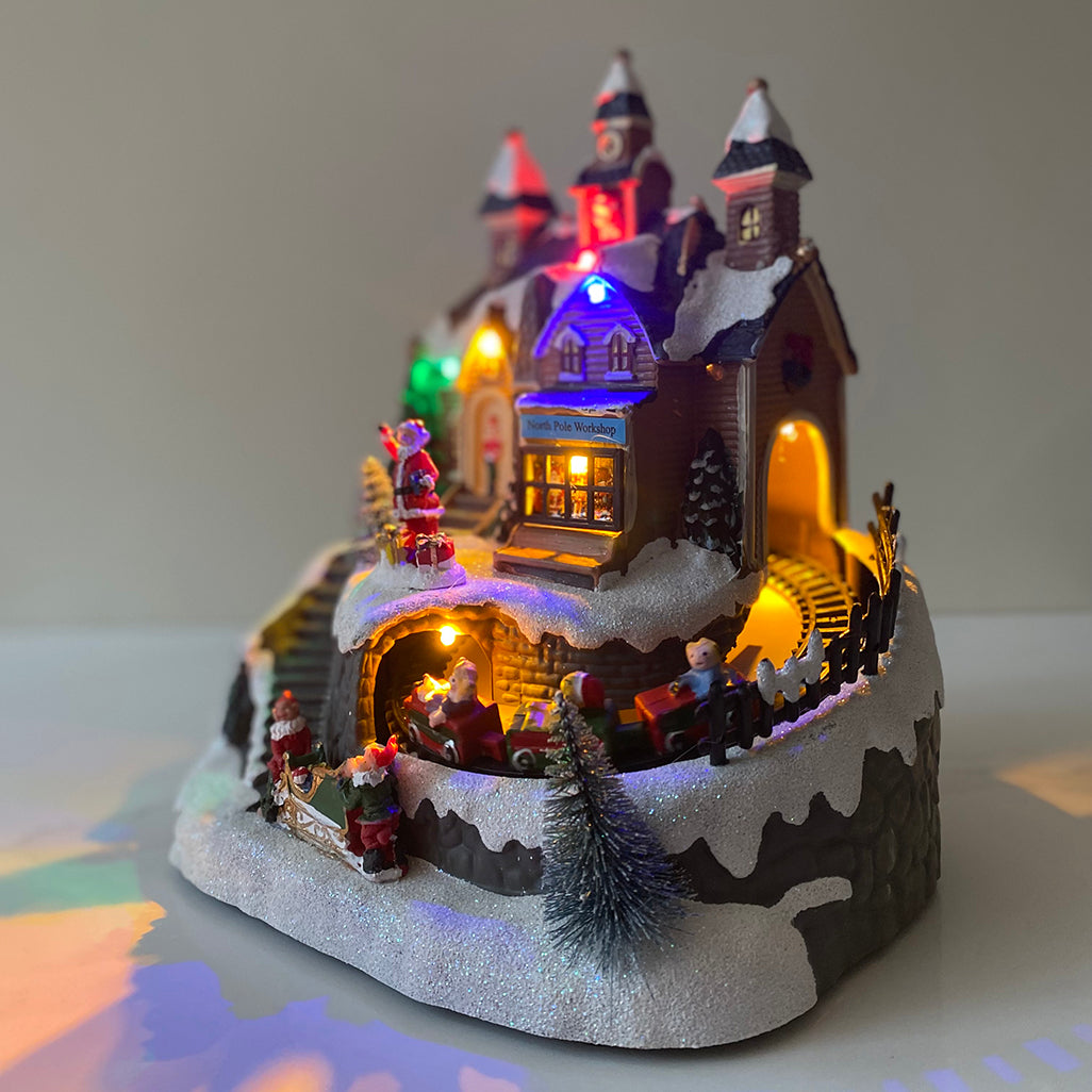 A festive Christmas scene ornament featuring Santa Claus, LED lighting, and a moving train with children passing through a tunnel. The decoration is designed for indoor use.