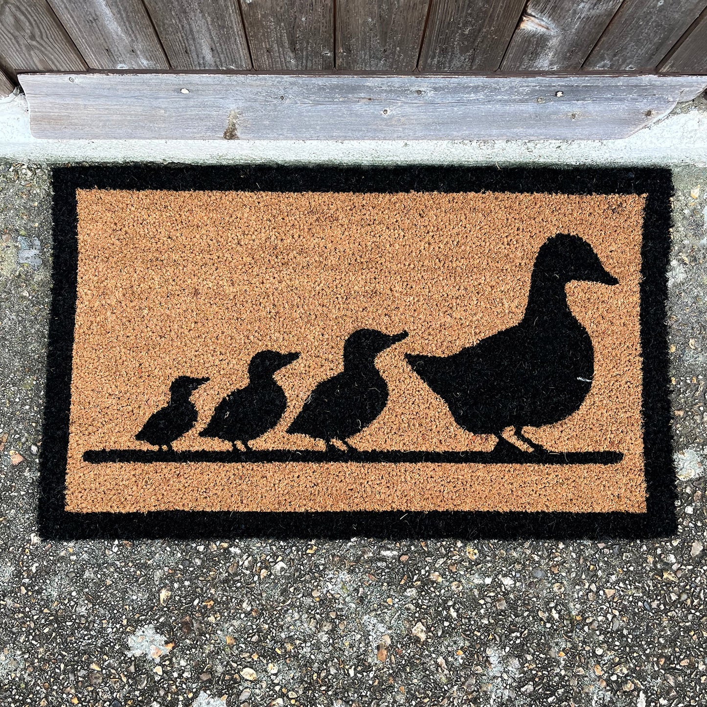 Rectangular beige coir doormat with a black silhouette of a duck family design, placed on a concrete surface.