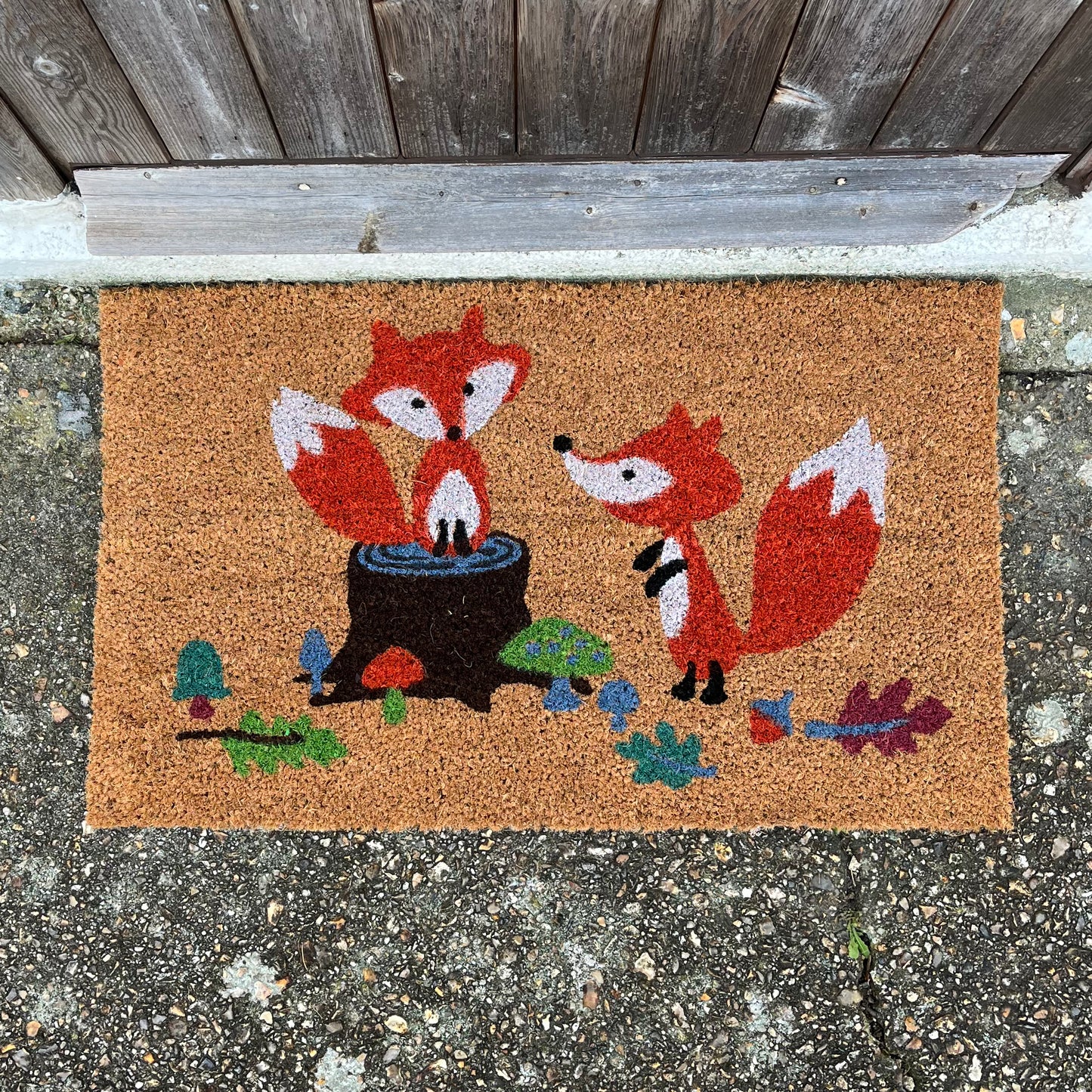 A coir doormat with an illustration of two foxes in a woodland glade, placed on a floor with a wooden door in the background.
