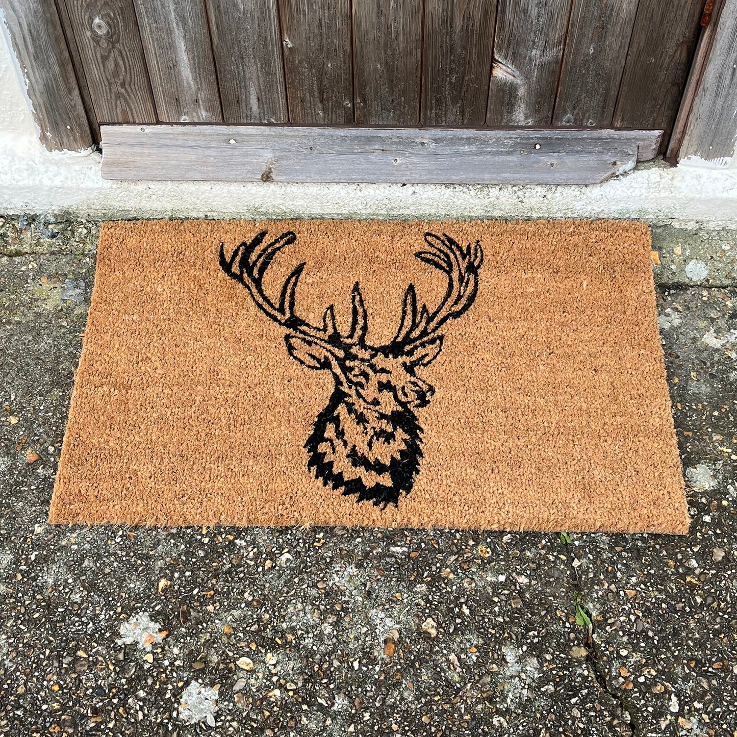 A rectangular beige coir doormat with a black silhouette of a stag's head design, placed on a concrete surface.