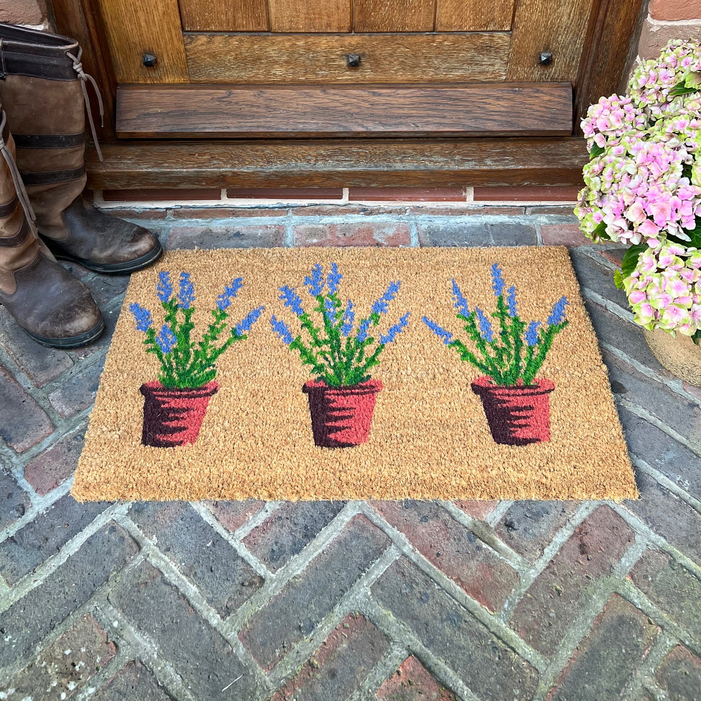 A coir doormat with an indoor and outdoor design, featuring three pots of lavender, placed on a doorstep.