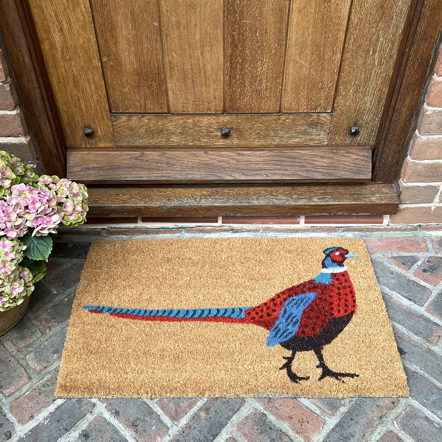 A coir doormat with a pheasant design placed on a doorstep, featuring a colorful bird illustration on a beige mat.