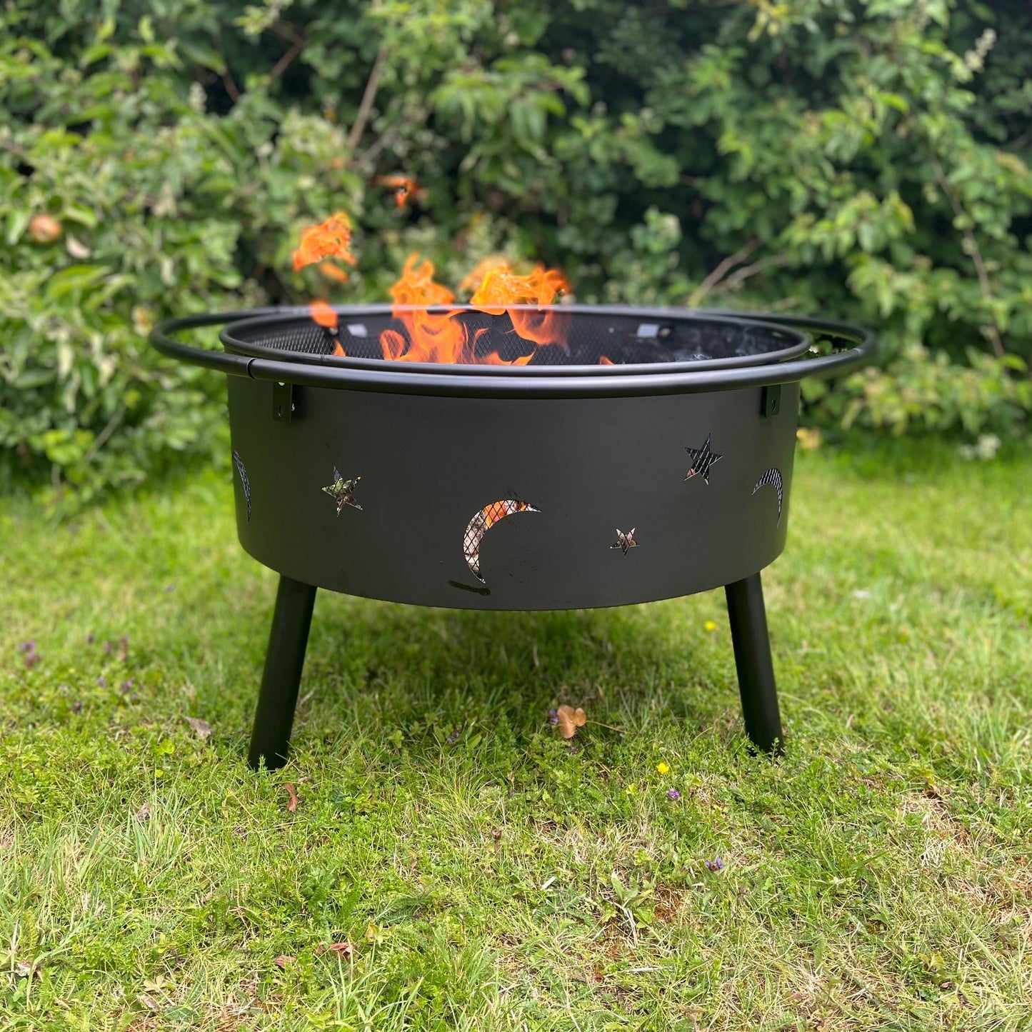 A black outdoor fire bowl with star and moon cut-outs and a spark guard, containing a fire that is burning brightly. The fire bowl is placed on a grassy surface.
