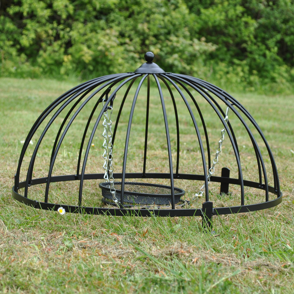 A black powder coated steel ground bird feeder cage with a dome shape and galvanised hanging chains, set on a grassy surface.