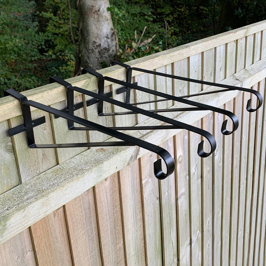 A set of four black painted steel hanging basket brackets installed on a wooden fence.
