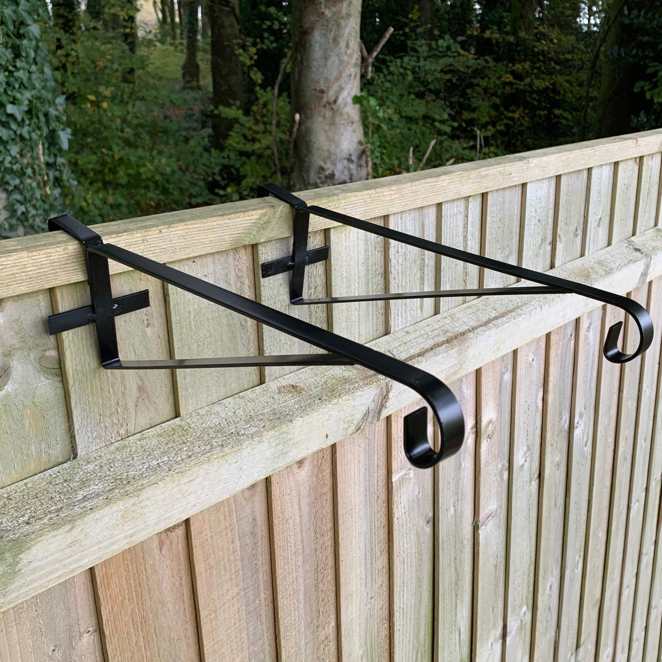 A set of two black painted steel hanging basket brackets installed on a wooden fence panel.