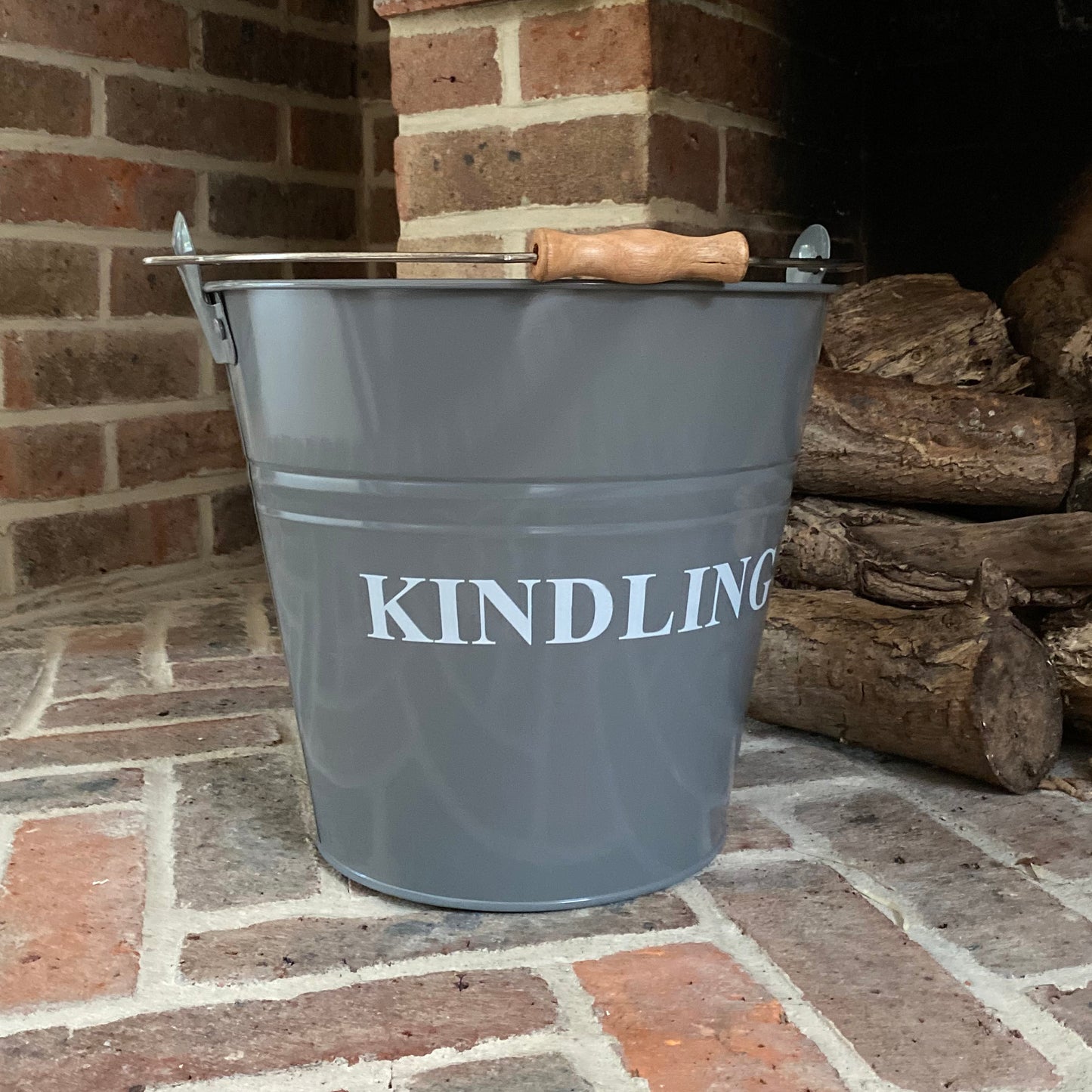 A French grey colored metal kindling bucket with a sturdy metal handle and an ergonomic wooden grip, placed on a brick hearth.