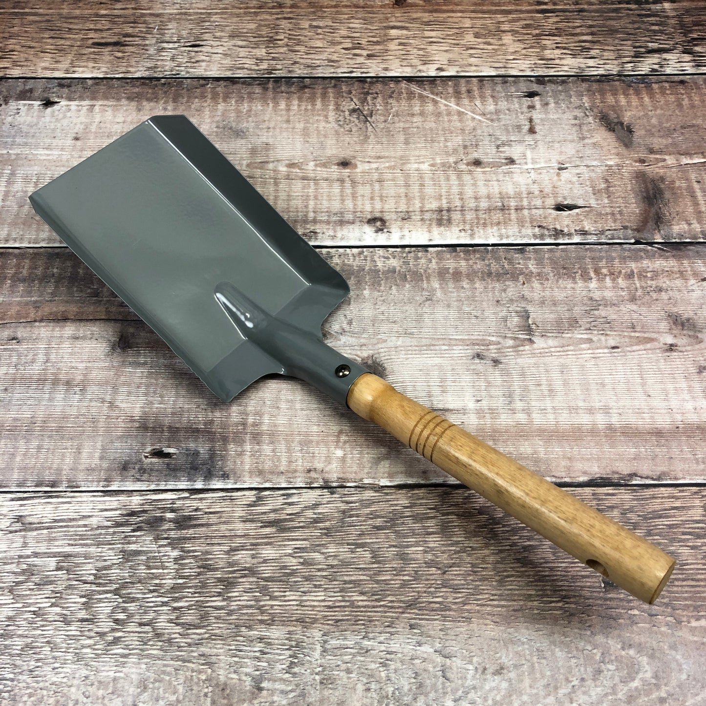 A French grey colored metal shovel with a wooden handle, placed on a wooden surface.