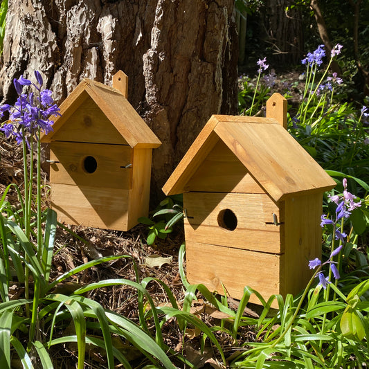 Two wooden multi-hole birdhouses placed in a garden setting with flowers and a tree in the background.