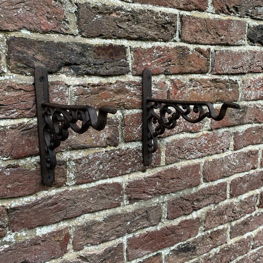 Two brown cast iron decorative hanging basket brackets mounted on a brick wall.