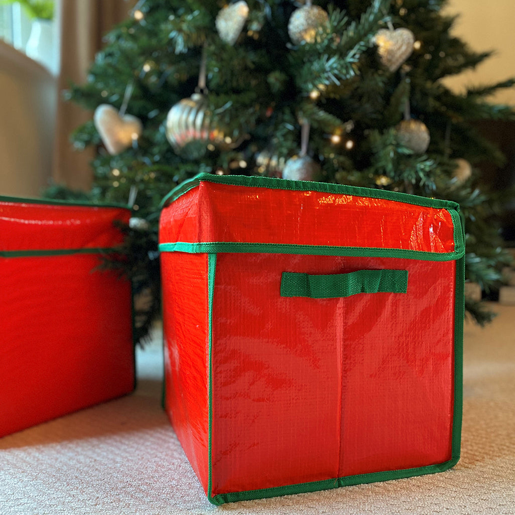 A red Christmas bauble decorations storage box with green trim, placed on a floor beside a decorated Christmas tree.