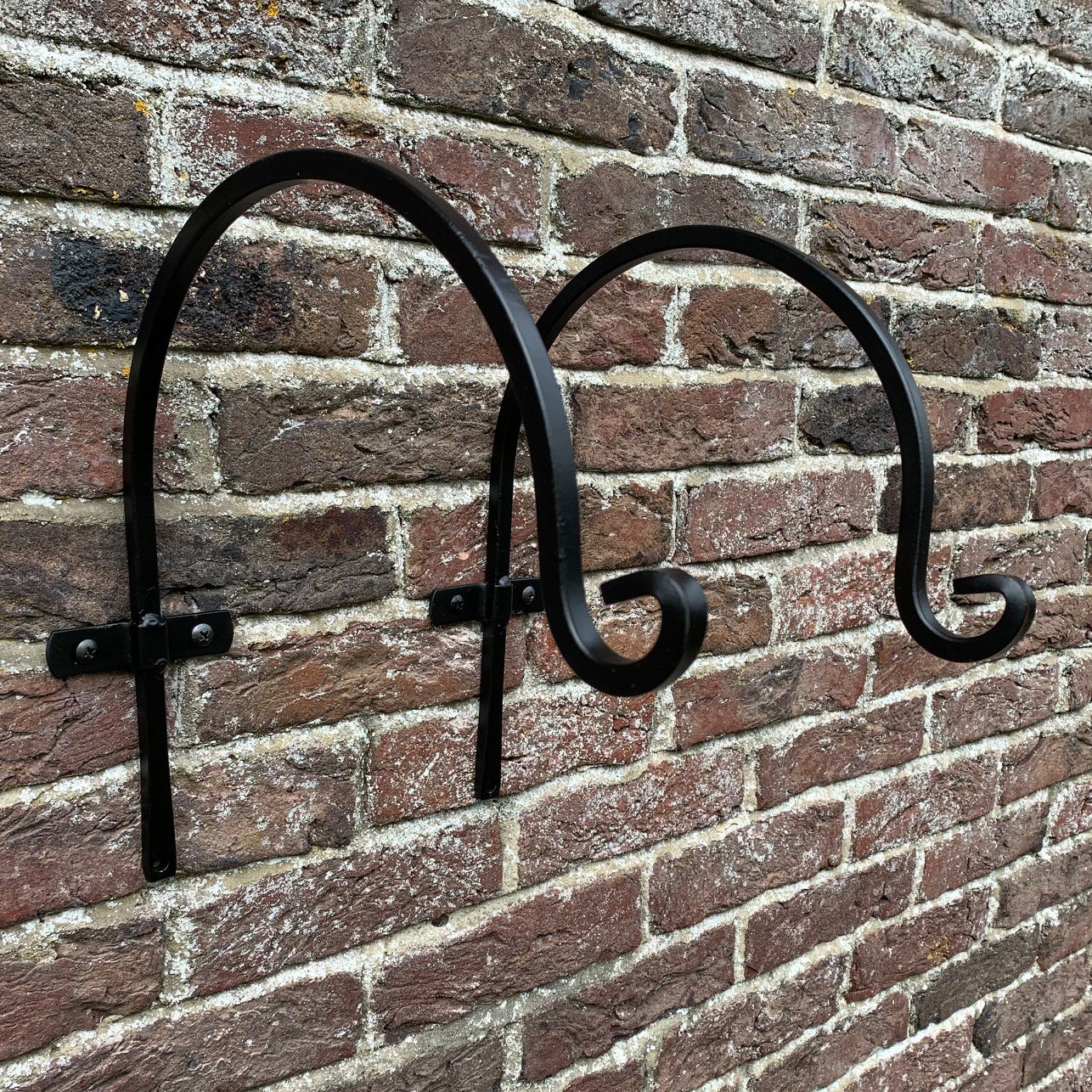Two black powder coated steel hooks designed for hanging baskets or planters, mounted on a brick wall.