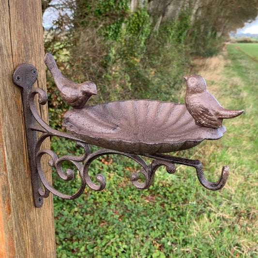 A wall-mounted cast iron bird bath feeder with two birds facing each other on the edges, and a floral bracket for hanging baskets.
