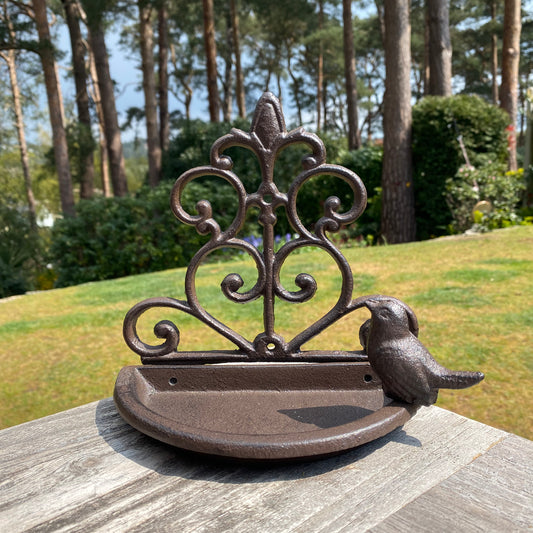 A brown cast iron wall mounted bird feeder with a decorative back panel and a bird perched on the rim, set against a grassy background.