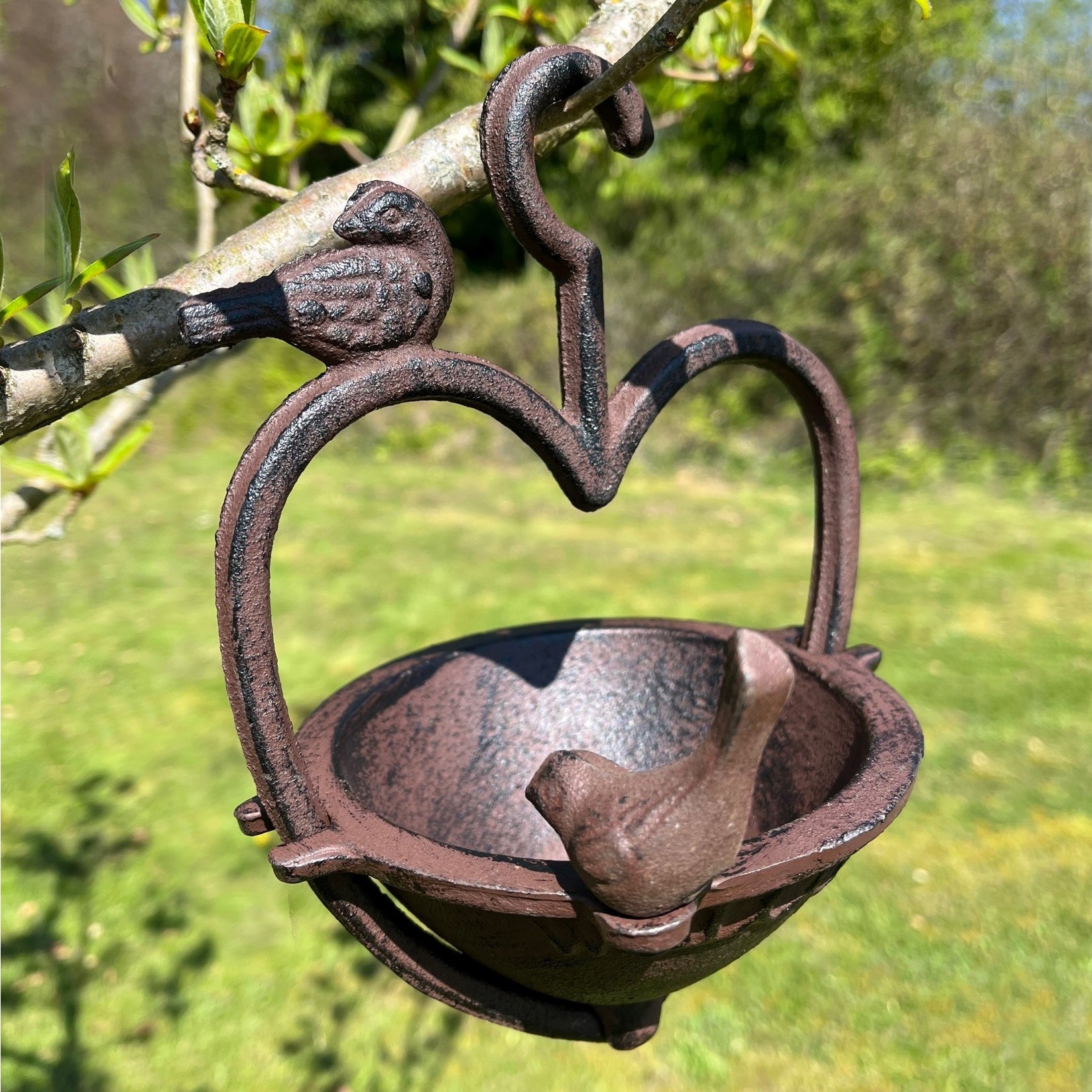 A heart-shaped cast iron bird feeder hanging from a branch with a bird perched on the rim, set against a blurred background of a garden.