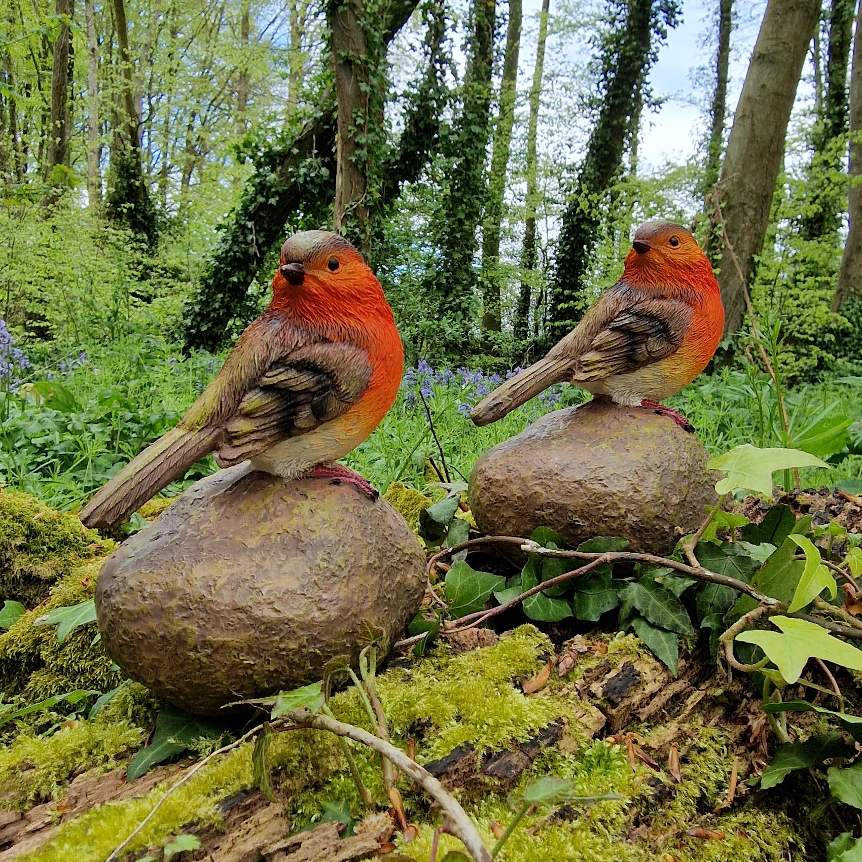 Two resin garden ornaments designed as Robins sitting on stones, placed in a natural outdoor setting with greenery around.