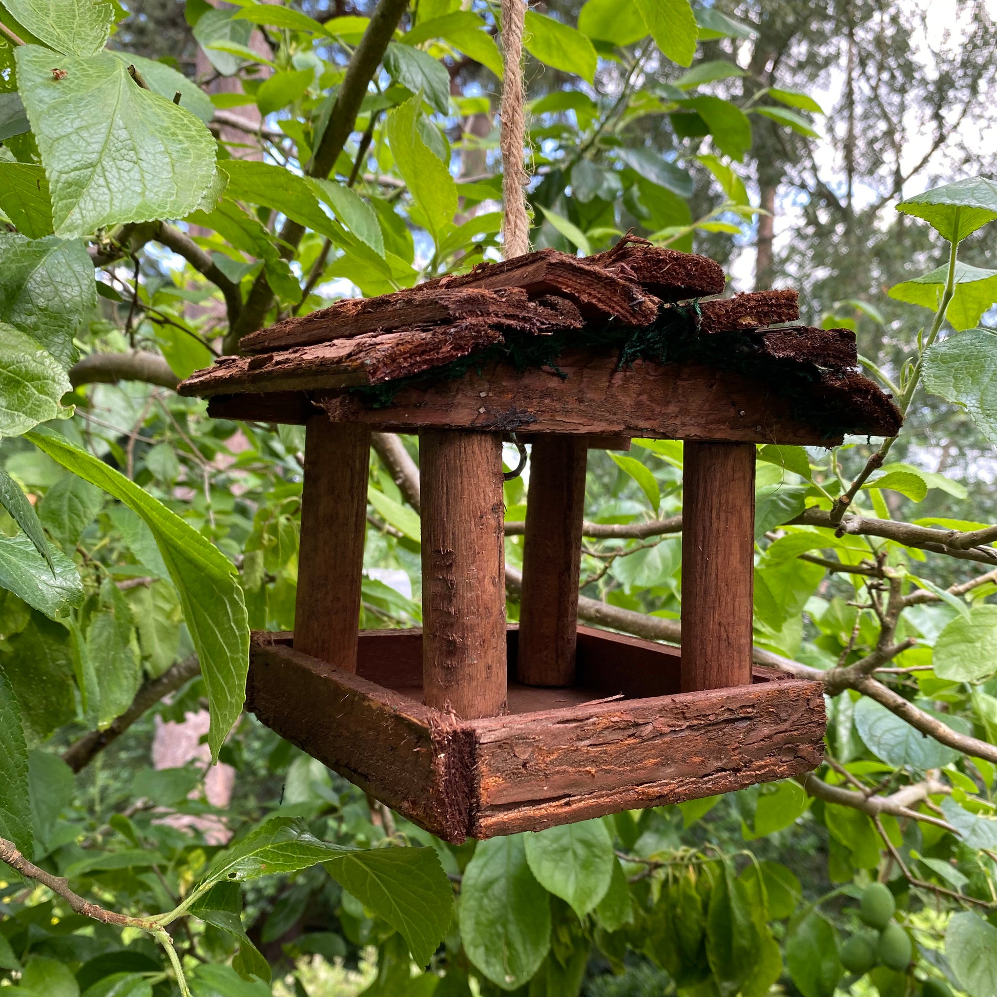 A rustic style hanging wooden bird table feeder placed among green foliage.