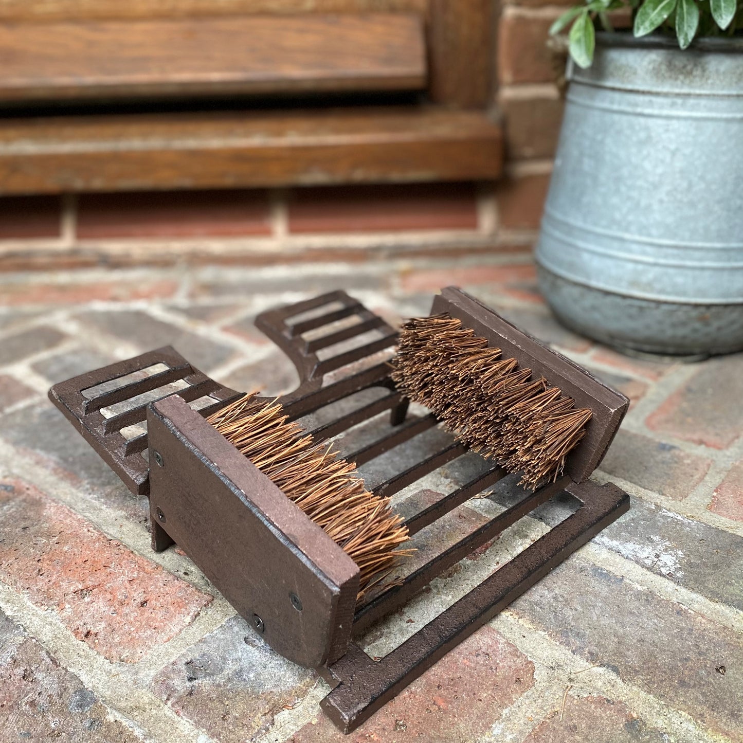 A traditional style cast iron boot jack and shoe scraper with bristle brushes, placed on a brick surface.