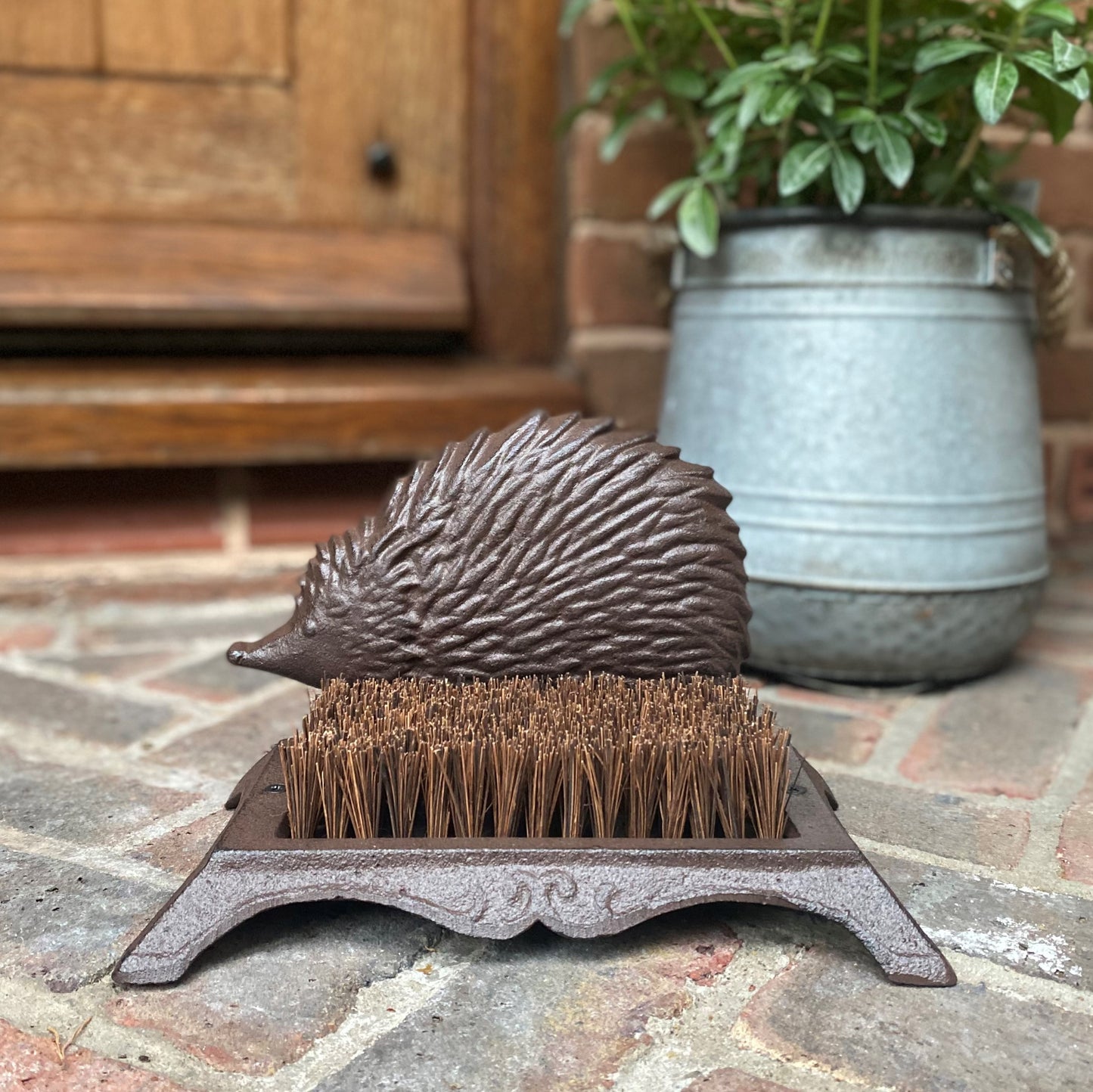 A vintage style cast iron boot brush shoe scraper with a hedgehog decoration on top, placed on a brick surface.