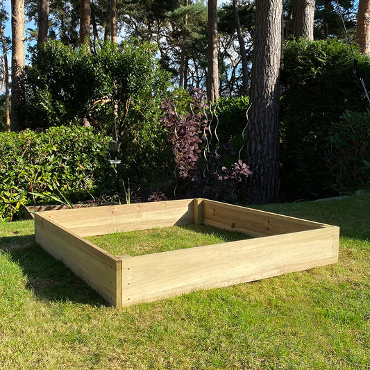 A wooden raised vegetable bed placed on grass with shrubs and trees in the background.