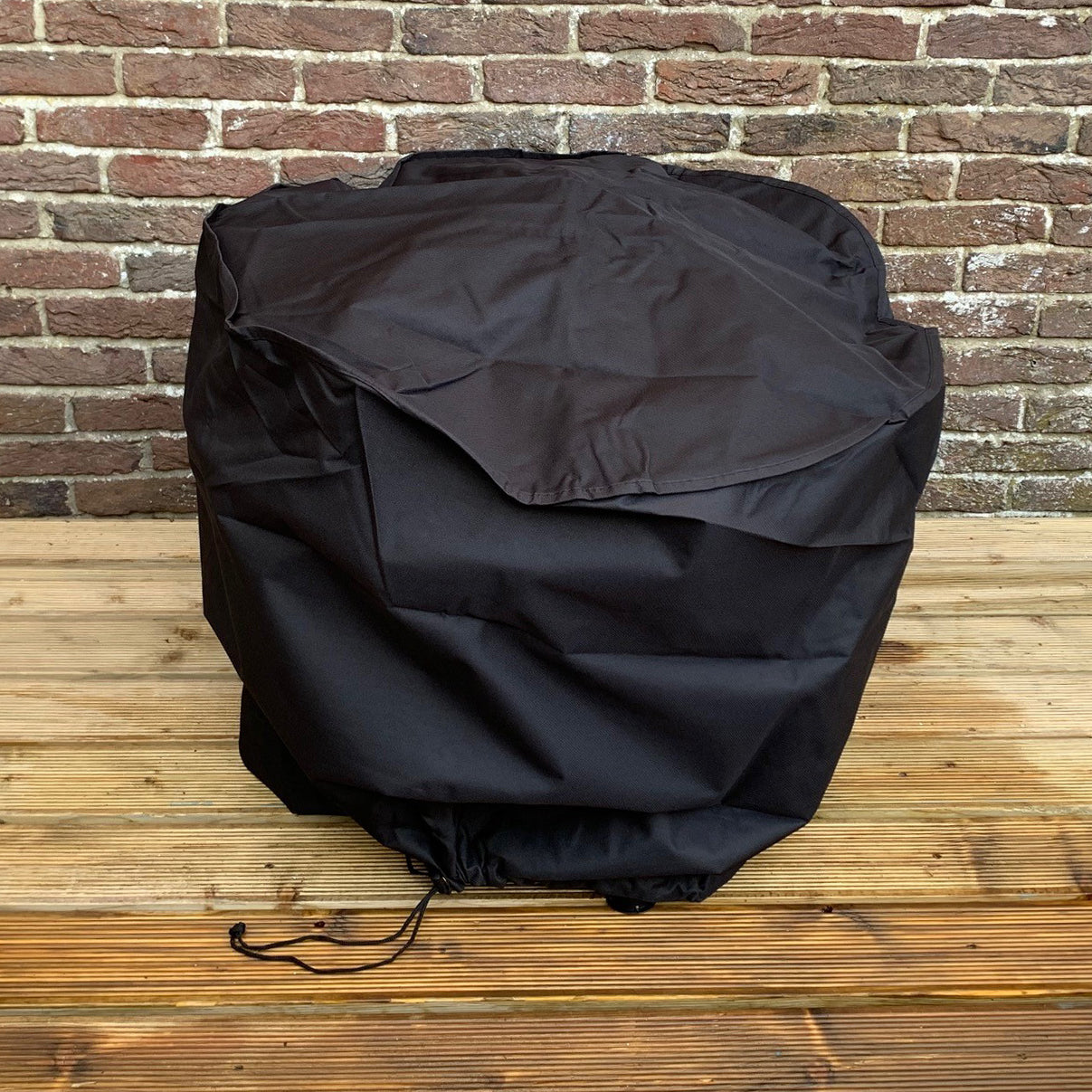 A black waterproof firepit cover placed on a wooden surface against a brick wall, featuring an adjustable fixing cord.