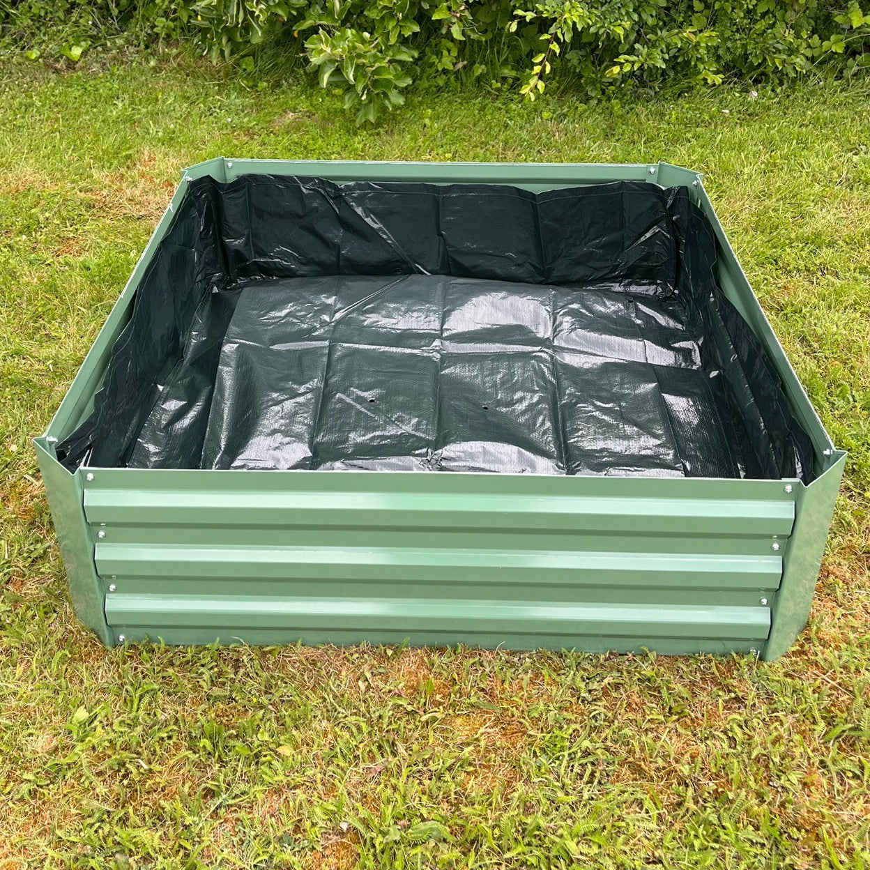 A black liner for a metal raised vegetable bed, placed inside the green bed which is set on a grass surface.