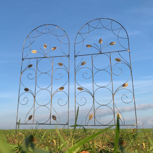 Two leaf design metal trellises placed upright on a grassy field against a blue sky.