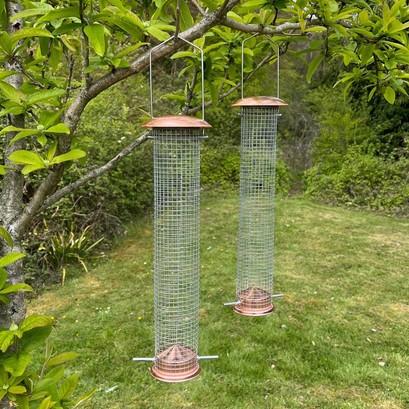 Two cylindrical copper-colored metal bird nut feeders with wire mesh, hanging from retractable hooks, set against a garden background.