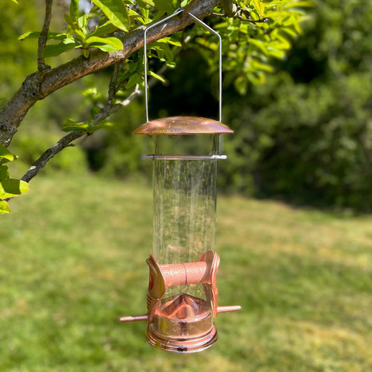 A copper style hanging bird seed feeder with two feeding ports, seen hanging from a retractable hook lid against a blurred green garden background.