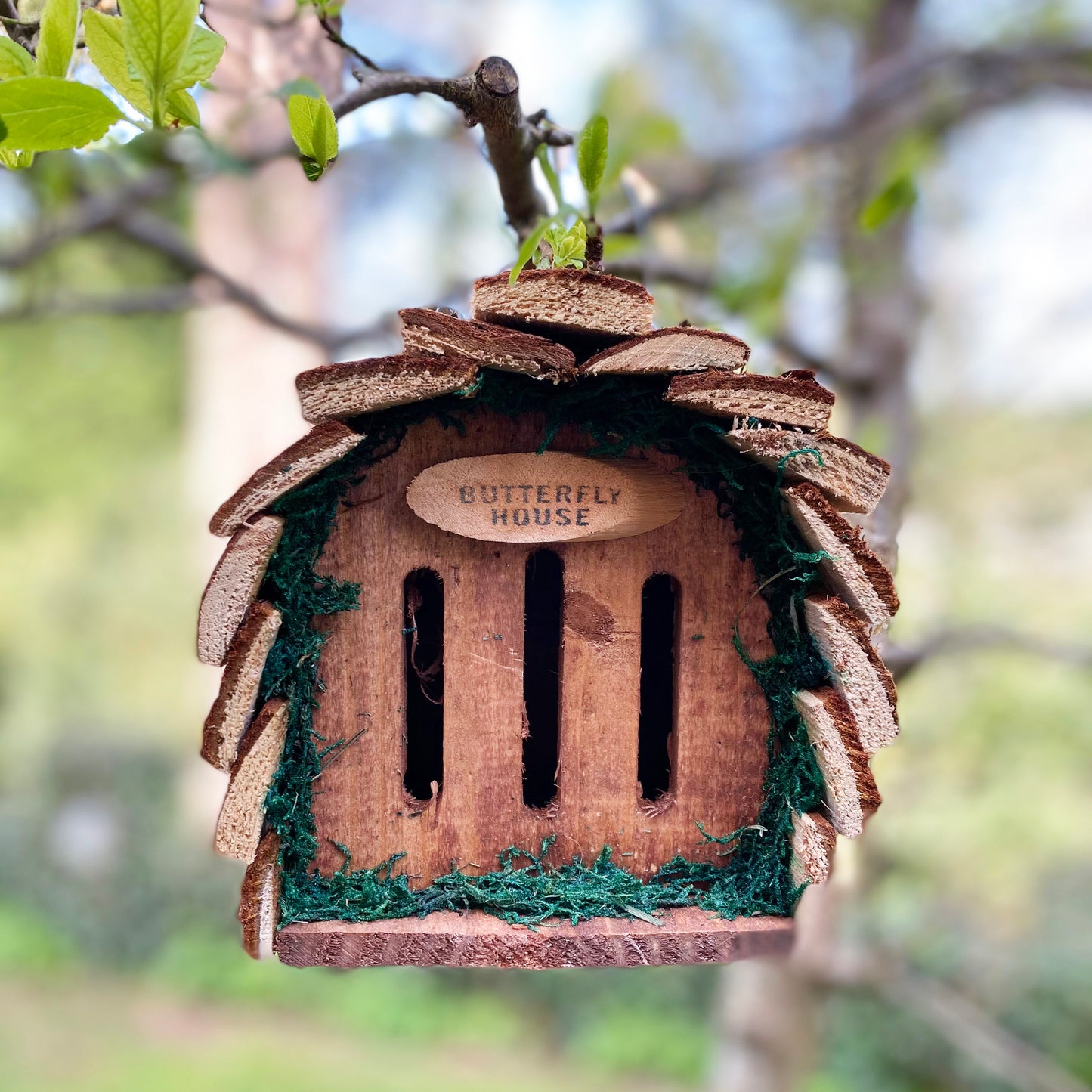 A rustic wooden butterfly house with a natural bark roof hanging from a metal hook, displayed against a blurred garden background.